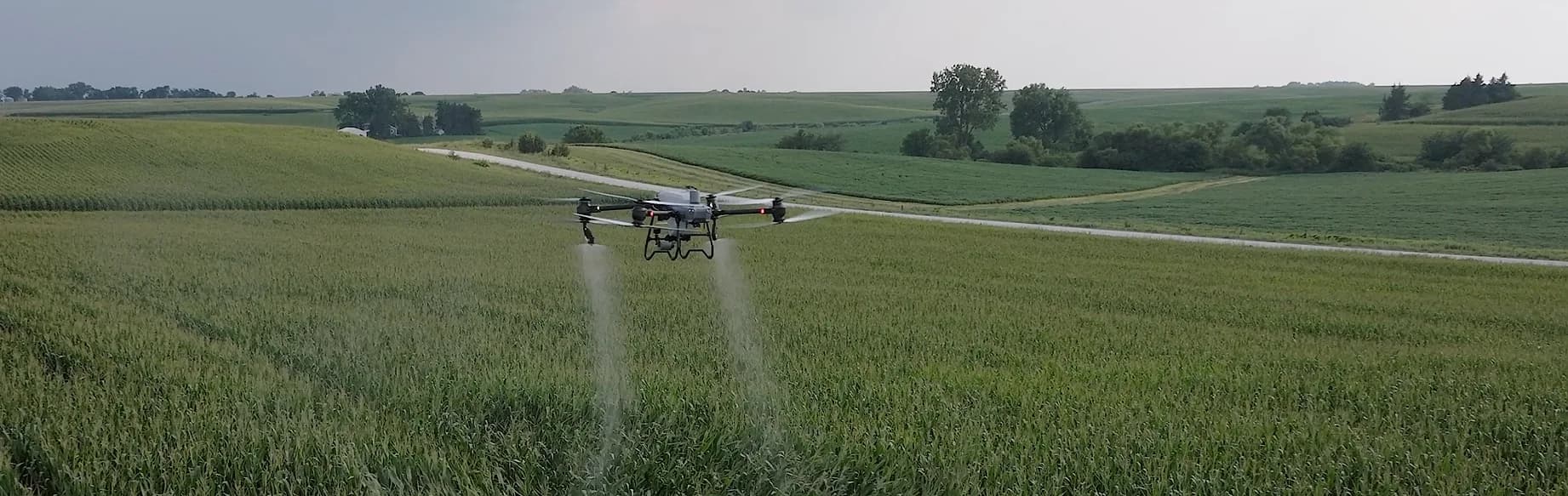 A spray drone mid-application over a green field, twin spray plumes visible behind it